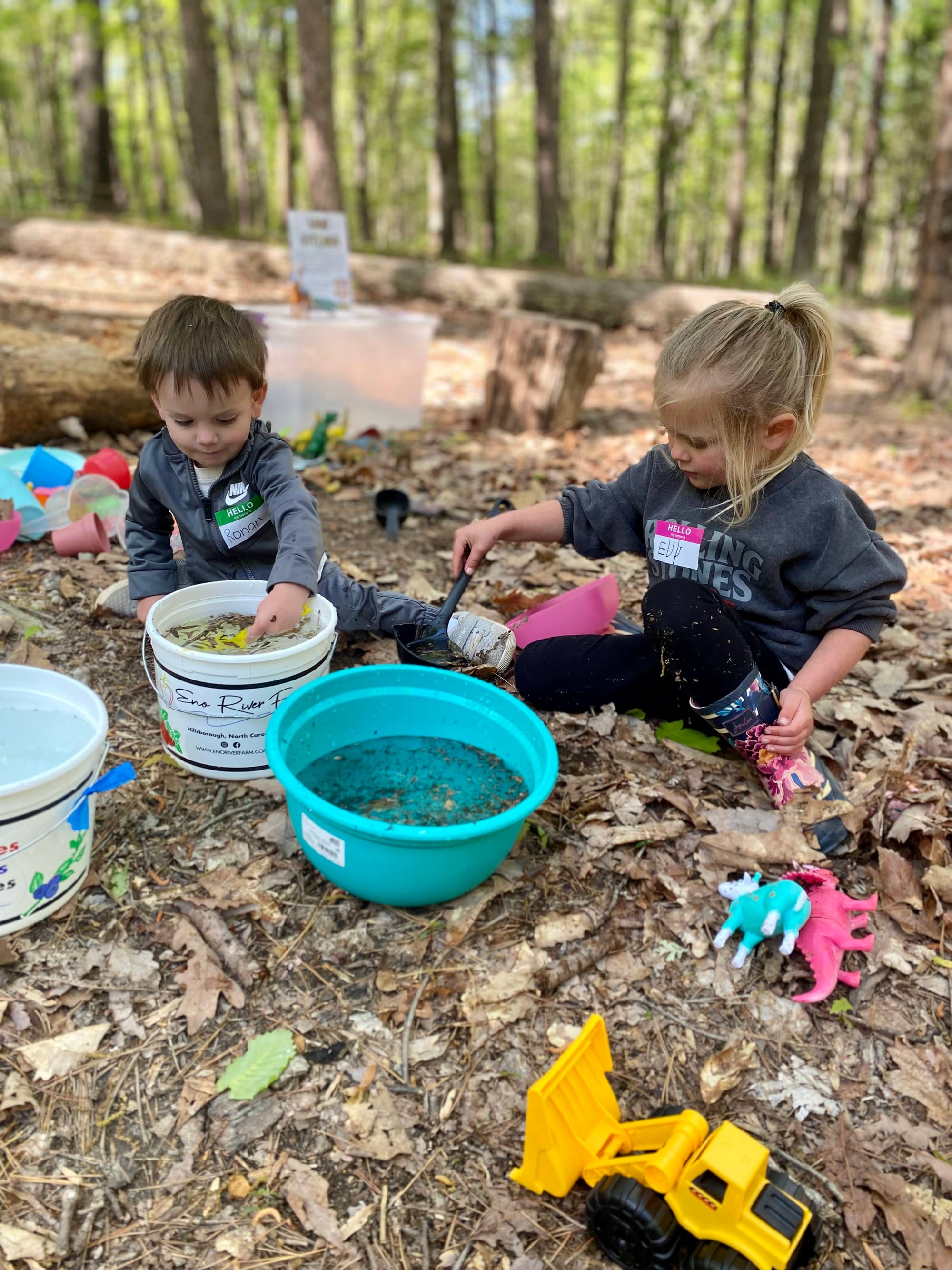 (Image: Two young children sitting in the leaves and dirty playing with buckets of water and scoopers with other toys scattered around them)