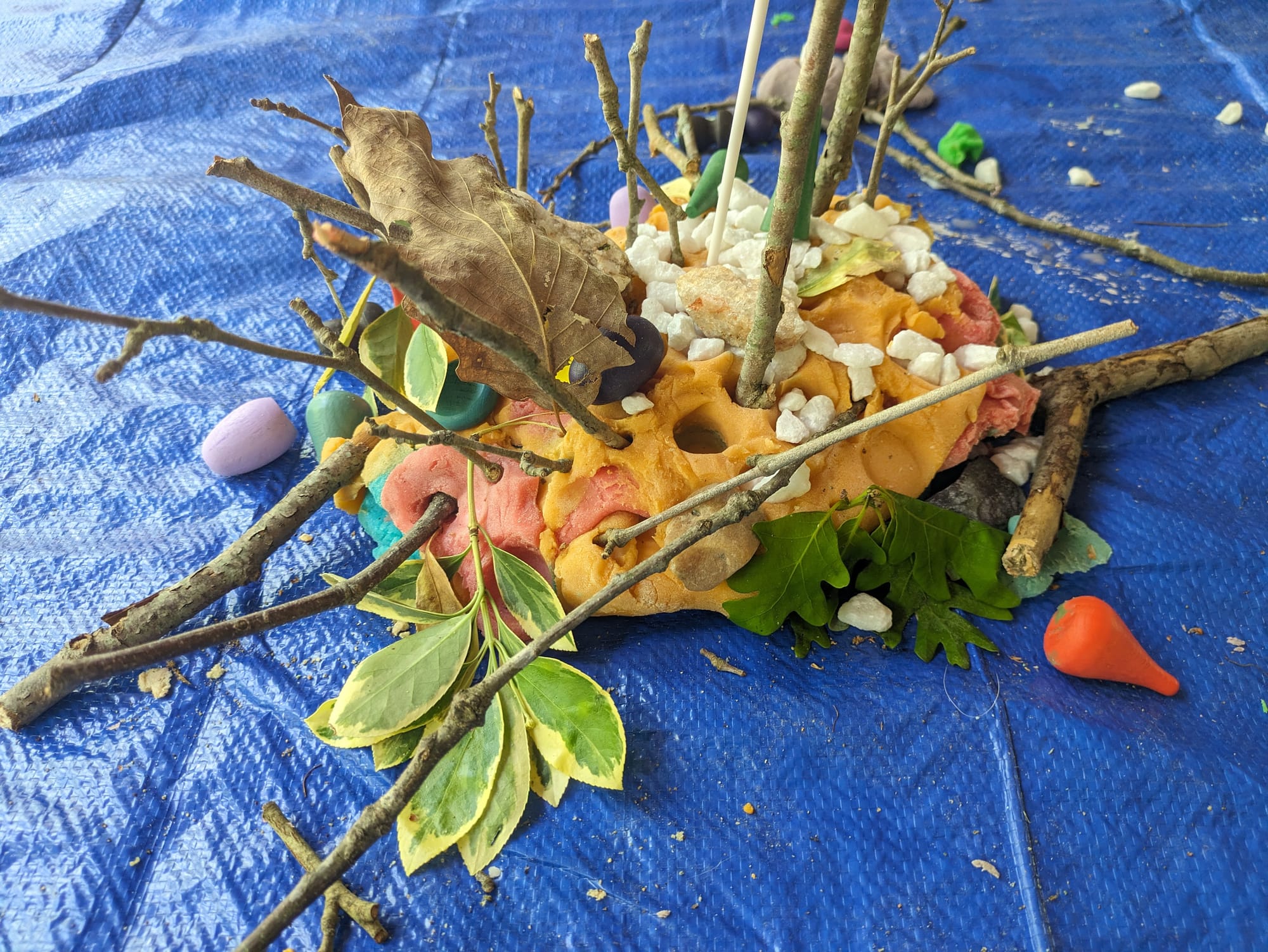 (Image: Close up photo of a large flat chunk of orange playdough with several sticks, rocks, leaves, and other little nature pieces sticking out of it in every direction)